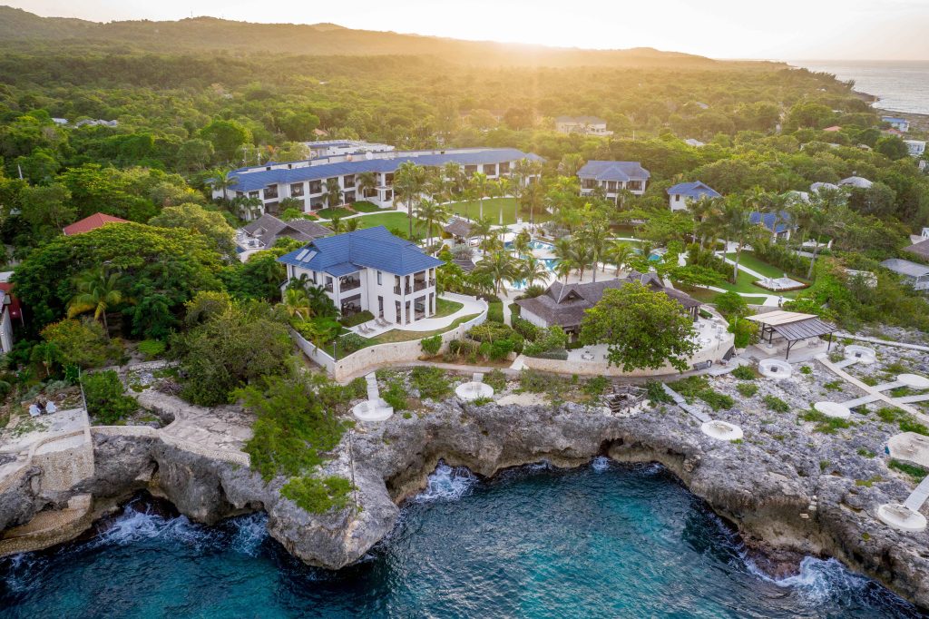 Aerial view of a tropical resort at sunrise, featuring lush greenery, swimming pools, and rocky coastline in Jamaica.