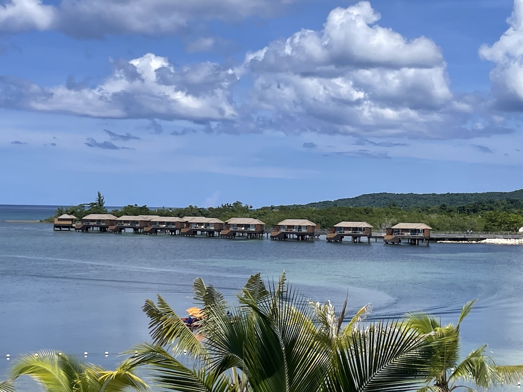 Overwater bungalows surrounded by calm blue waters and lush greenery in Jamaica under a partly cloudy sky.