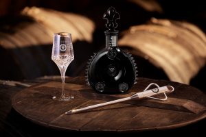A black bottle of LOUIS XIII Rare Cask 42.1 sits beside an elegant crystal glass and a tasting tool on a wooden table.