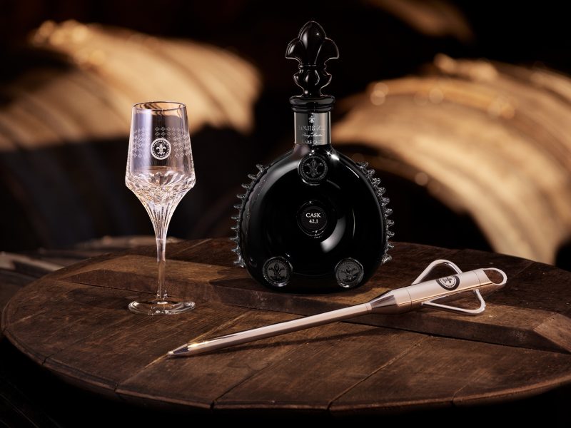 A black bottle of LOUIS XIII Rare Cask 42.1 sits beside an elegant crystal glass and a tasting tool on a wooden table.