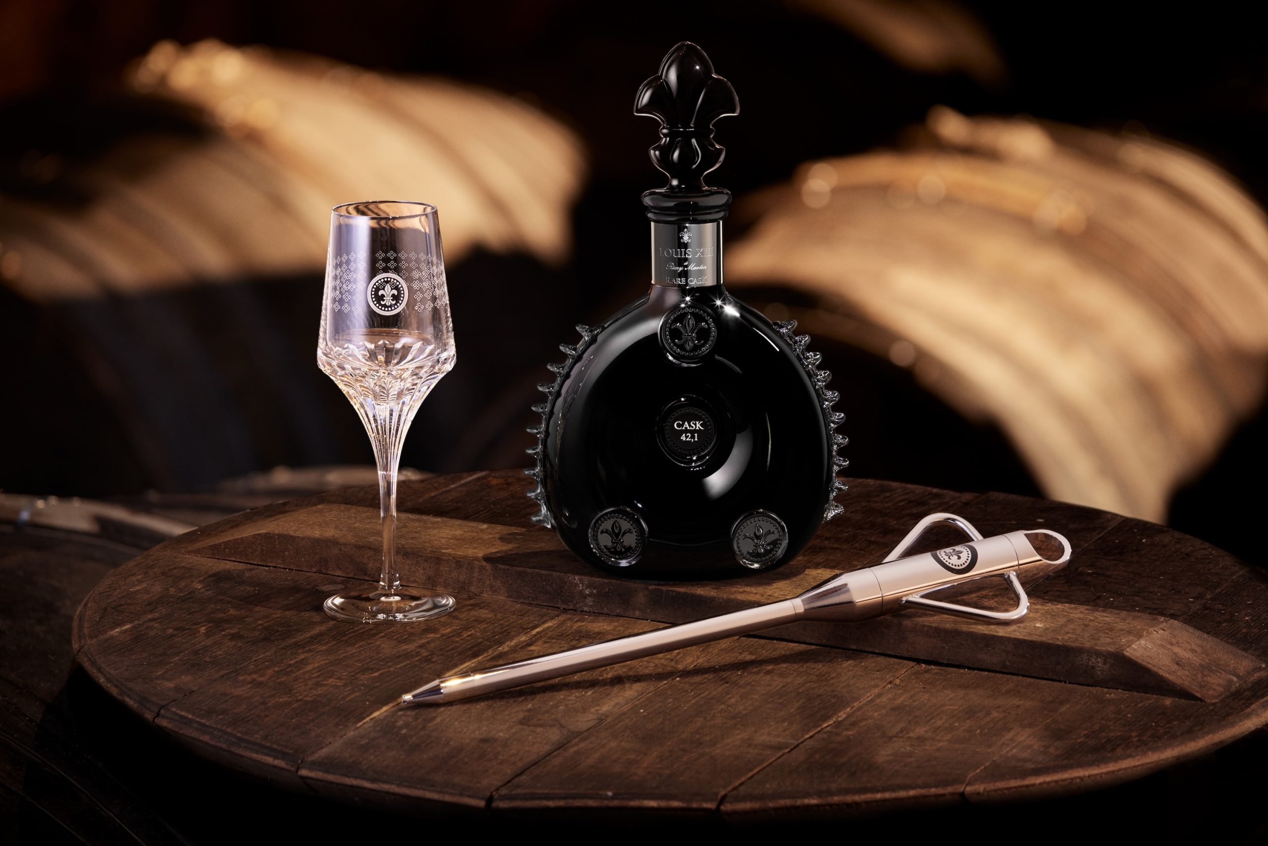 A black bottle of LOUIS XIII Rare Cask 42.1 sits beside an elegant crystal glass and a tasting tool on a wooden table.