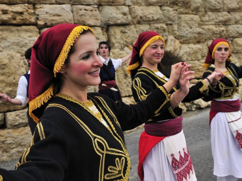 Traditional Cretan dancers perform in traditional attire, showcasing the vibrant culture of Crete against a stone backdrop.