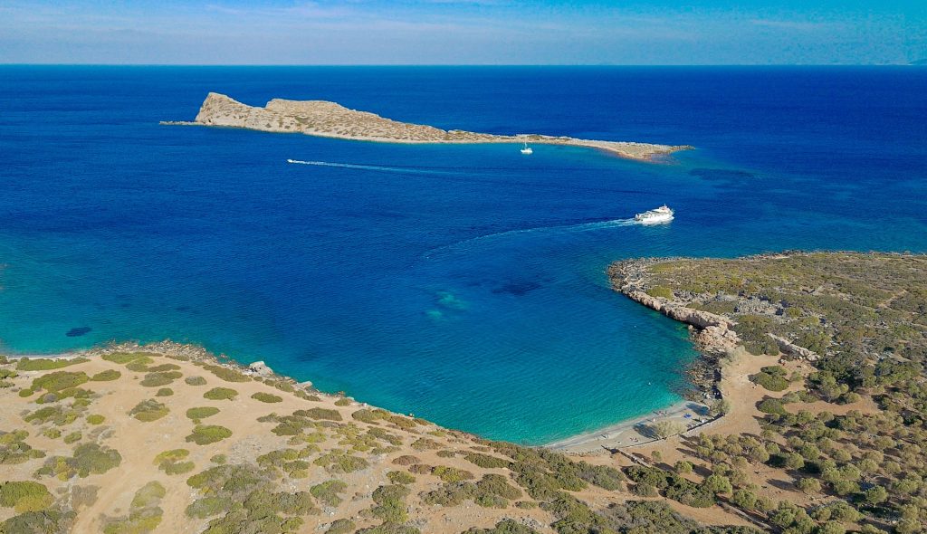Aerial view of a serene blue sea with boats, rugged coastline, and sandy beach surrounded by greenery in Crete.