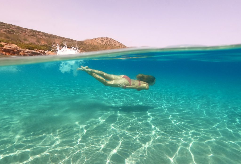 A person swimming underwater in clear turquoise water, with sunlight creating patterns on the sandy bottom.