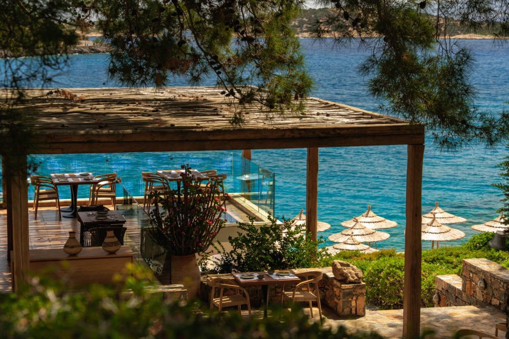 Terrace dining area overlooking the turquoise sea, with tables set among greenery and beach umbrellas in the background.