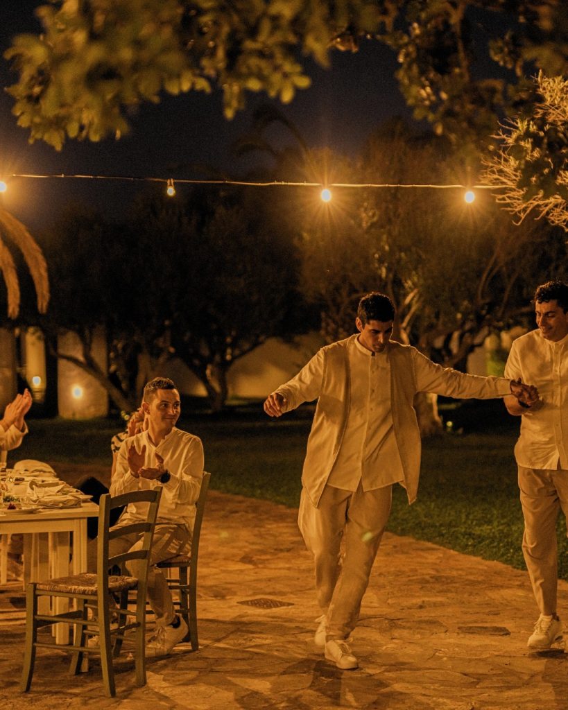 A group celebrates outdoors at night, enjoying dinner and dancing under string lights in a scenic garden setting.