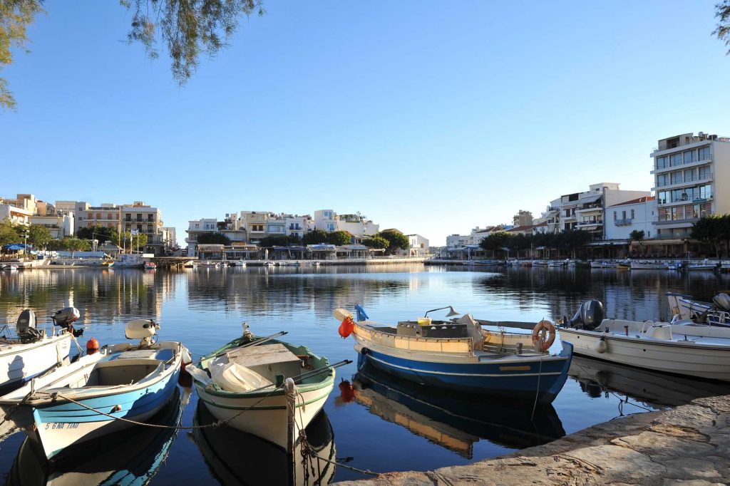 Harbor scene in Crete with colorful boats moored and calm water reflecting nearby buildings under a clear blue sky.