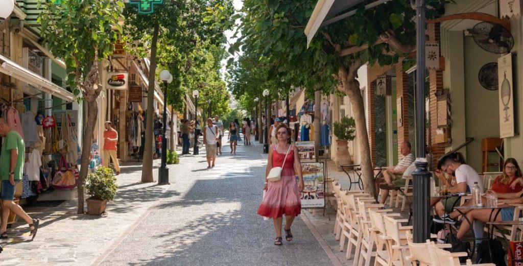 A bustling street in Crete lined with shops and cafes, featuring pedestrians, trees, and a woman in a pink dress walking.