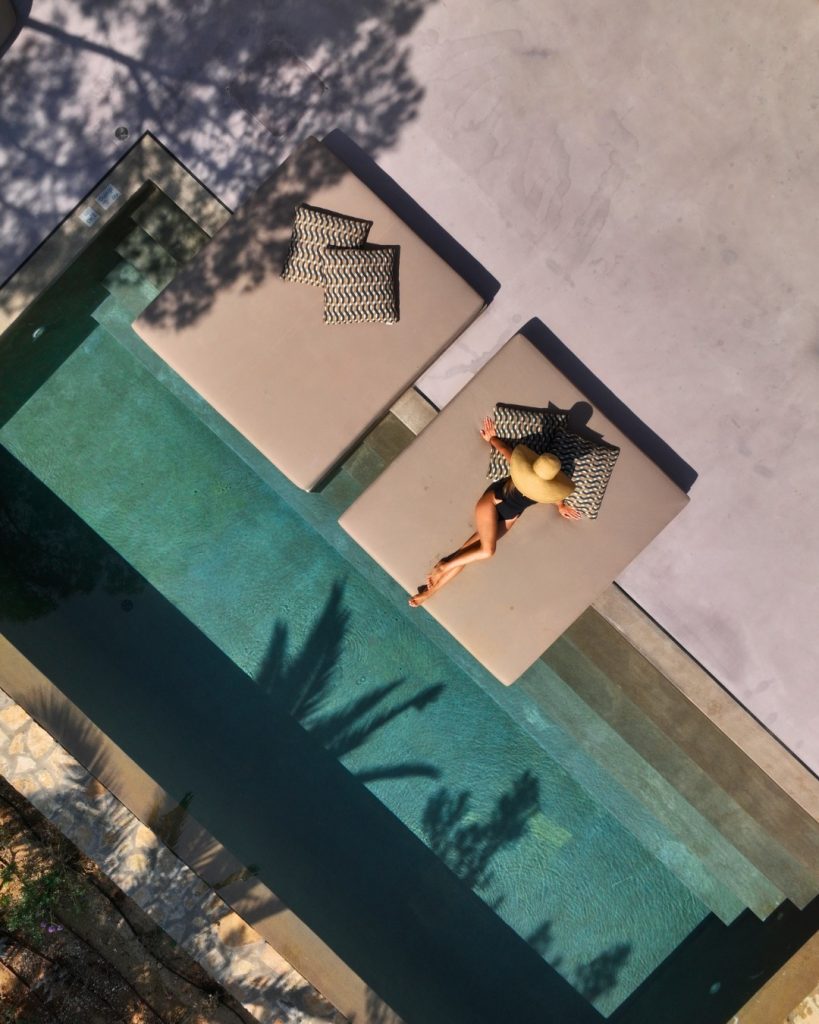 Aerial view of a woman relaxing on a poolside lounger, wearing a wide-brimmed hat and surrounded by turquoise water.