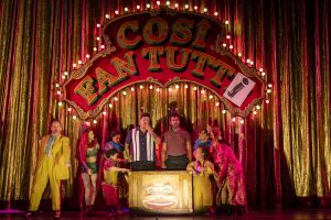 The cast performs under a vibrant sign for Così fan tutte, showcasing colorful costumes and a lively theatrical atmosphere.