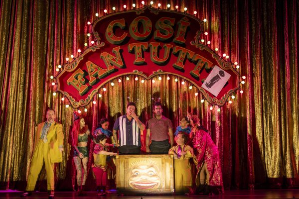 The cast performs under a vibrant sign for Così fan tutte, showcasing colorful costumes and a lively theatrical atmosphere.