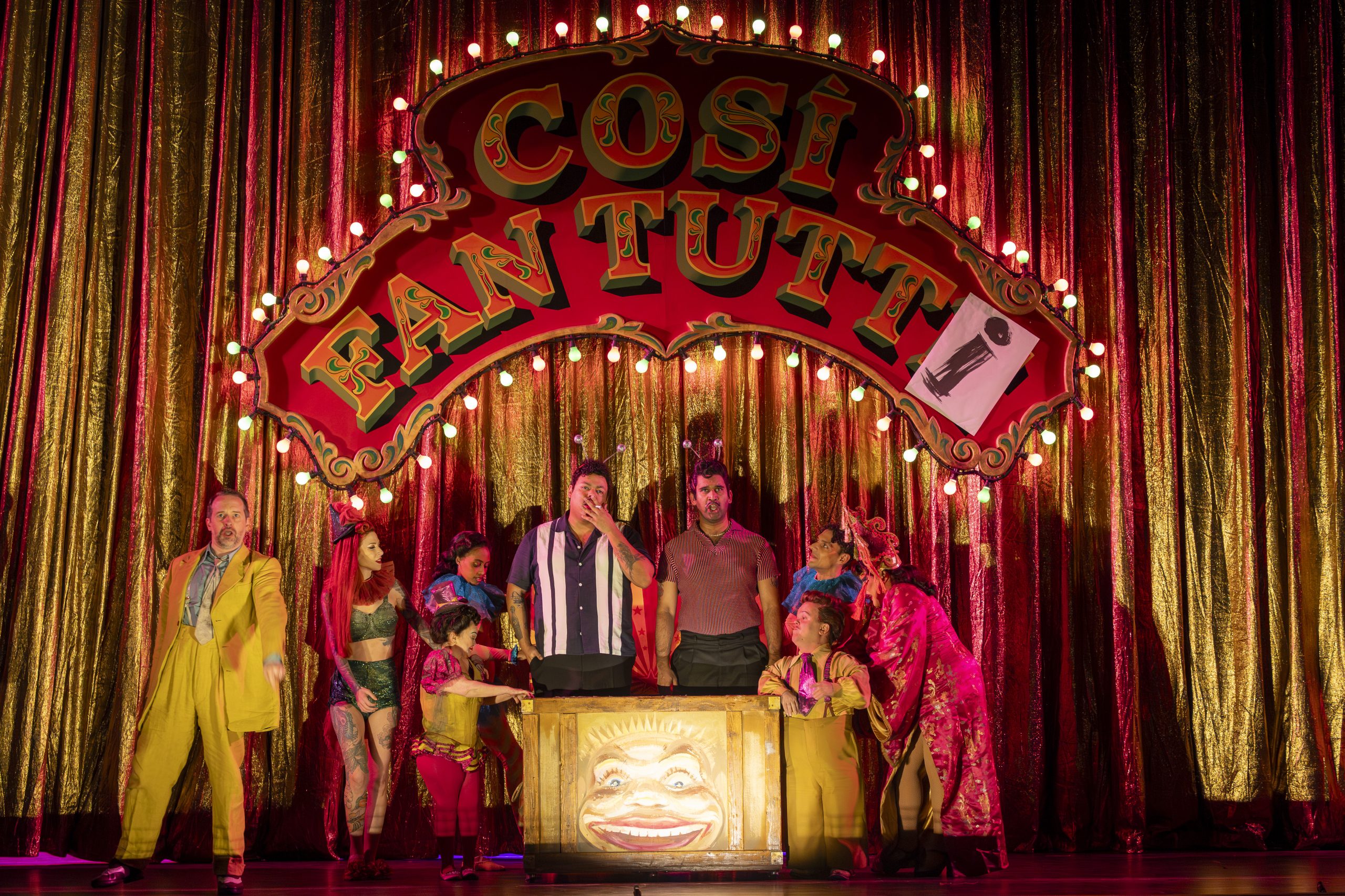 The cast performs under a vibrant sign for Così fan tutte, showcasing colorful costumes and a lively theatrical atmosphere.
