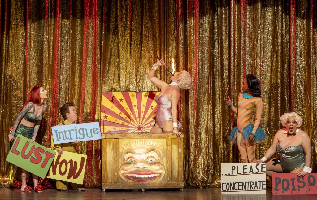 Performers in vibrant costumes hold signs with playful phrases during ENO’s Così fan tutte at London Coliseum.