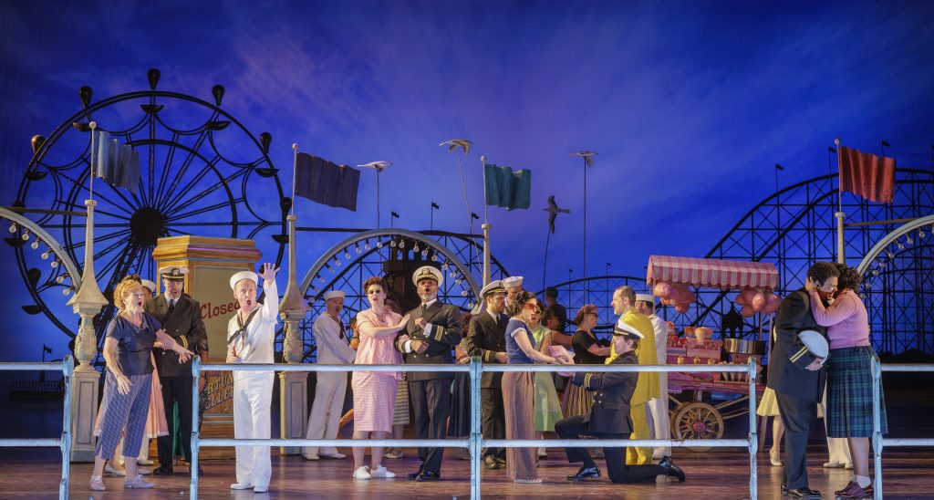 A lively performance scene from ENO's Così fan tutte, featuring a colorful fairground backdrop and engaged characters.
