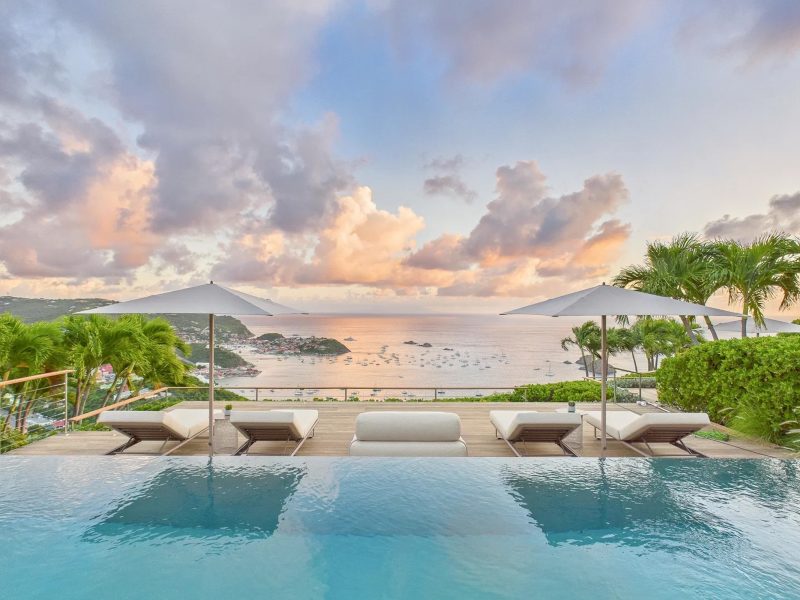 Infinity pool overlooking the ocean at a luxury villa, with lounge chairs and palm trees under a colorful sunset sky.