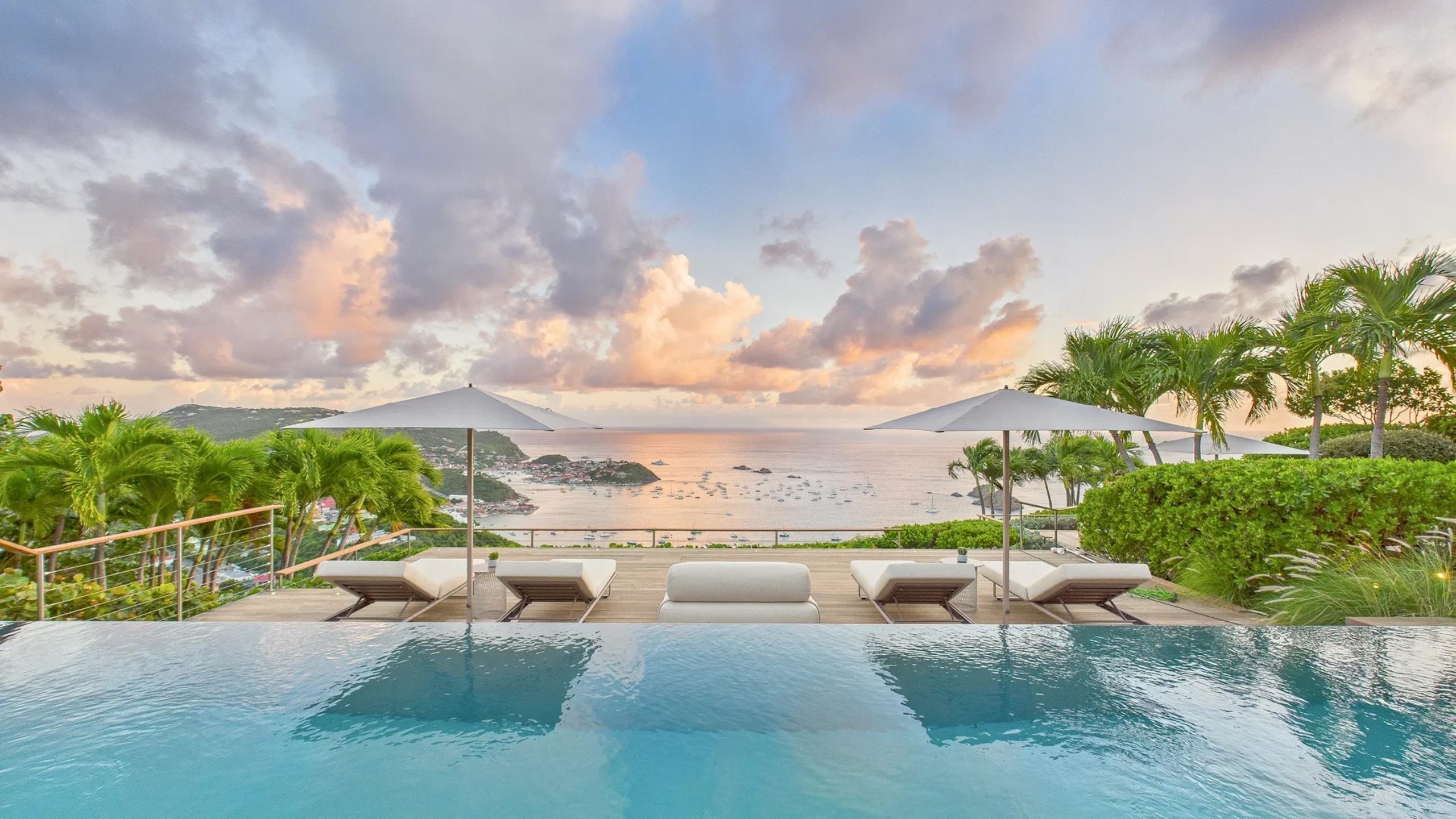 Infinity pool overlooking the ocean at a luxury villa, with lounge chairs and palm trees under a colorful sunset sky.