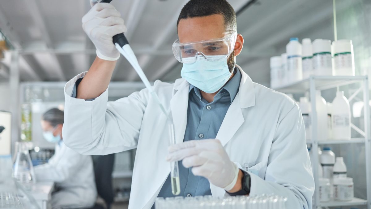 Scientist in a lab coats using a pipette to transfer liquid into a test tube, with laboratory equipment in the background.