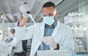 Scientist in a lab coats using a pipette to transfer liquid into a test tube, with laboratory equipment in the background.