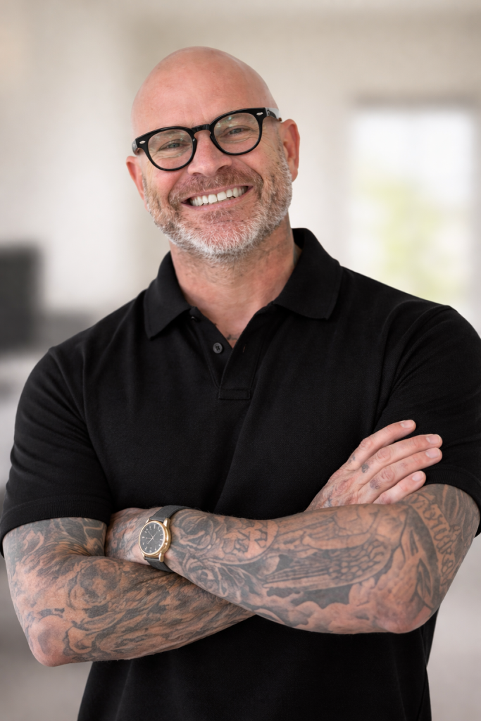 James Shipley, co-founder of PEARL, smiles confidently with arms crossed, showcasing his tattoos and casual black polo shirt.