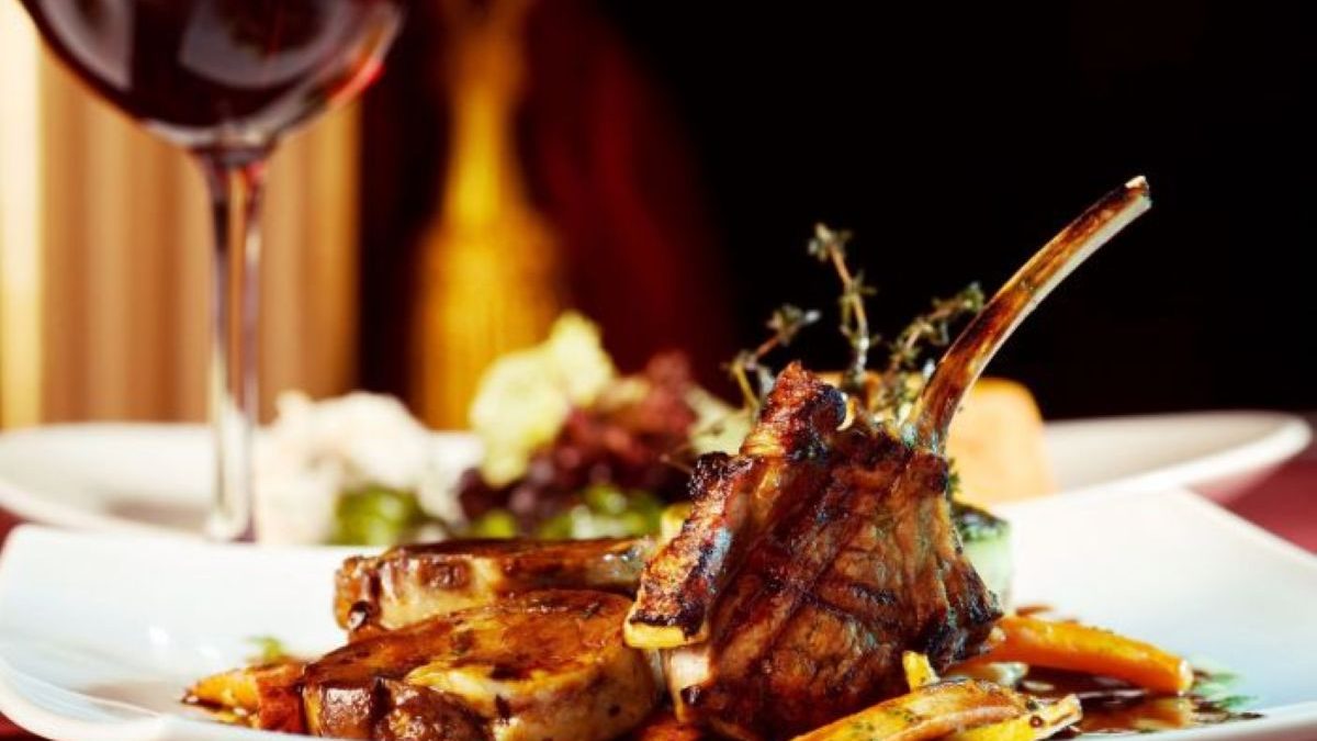 Grilled lamb chops served with vegetables and a glass of red wine in an upscale dining setting.