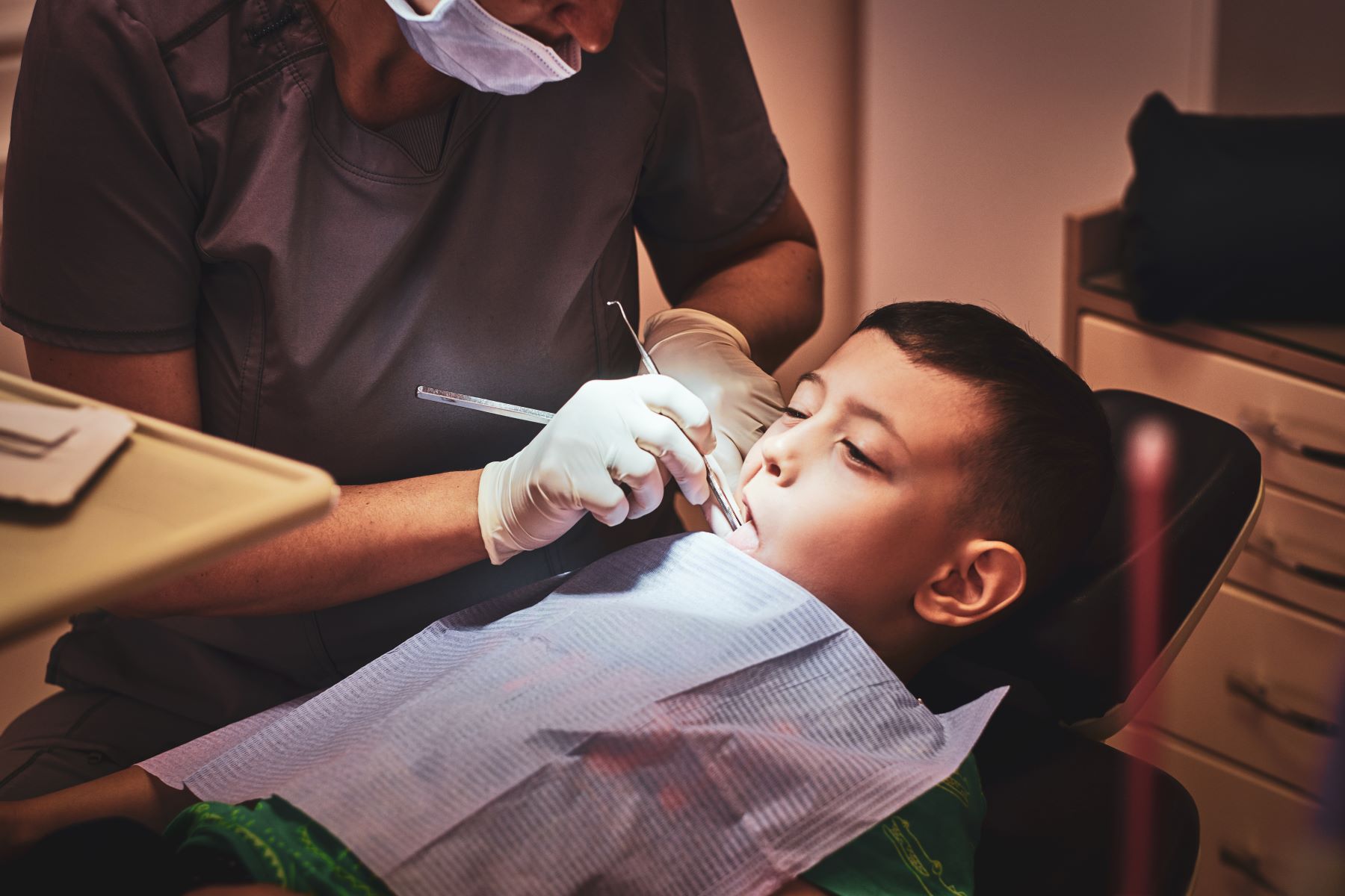A dentist examines a child's teeth using a dental tool while the child sits in a dental chair.