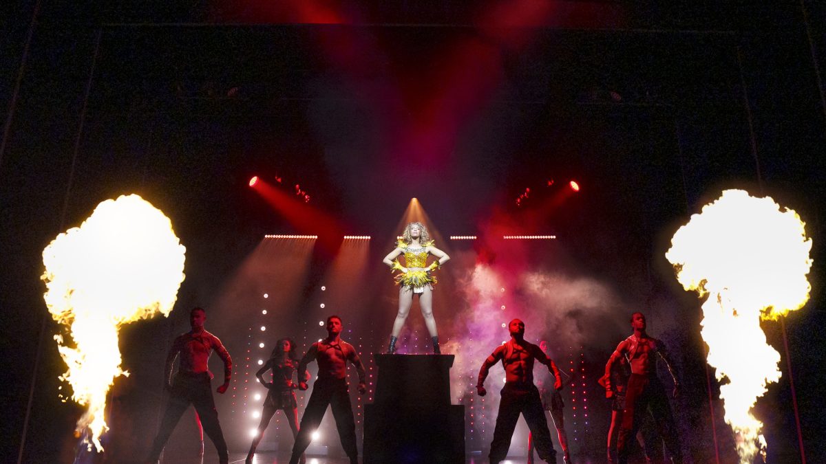 A performer in a vibrant costume stands on a pedestal surrounded by dancers and fire effects on stage.