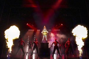 A performer in a vibrant costume stands on a pedestal surrounded by dancers and fire effects on stage.