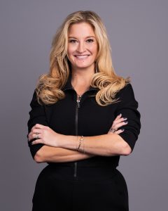 Woman with long blonde hair smiling confidently, wearing a black zip-up top and crossing her arms, against a gray background.