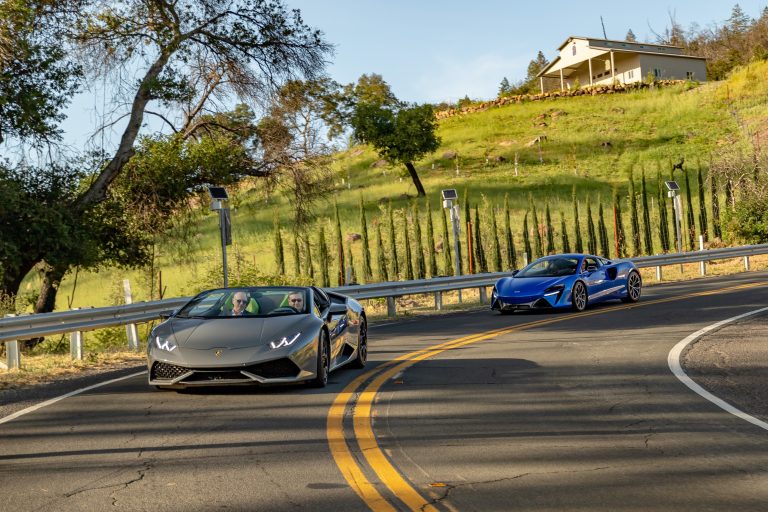 Luxury sports cars cruising on a winding road in Napa Valley, surrounded by lush greenery and scenic views.