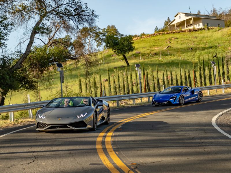 Luxury sports cars cruising on a winding road in Napa Valley, surrounded by lush greenery and scenic views.