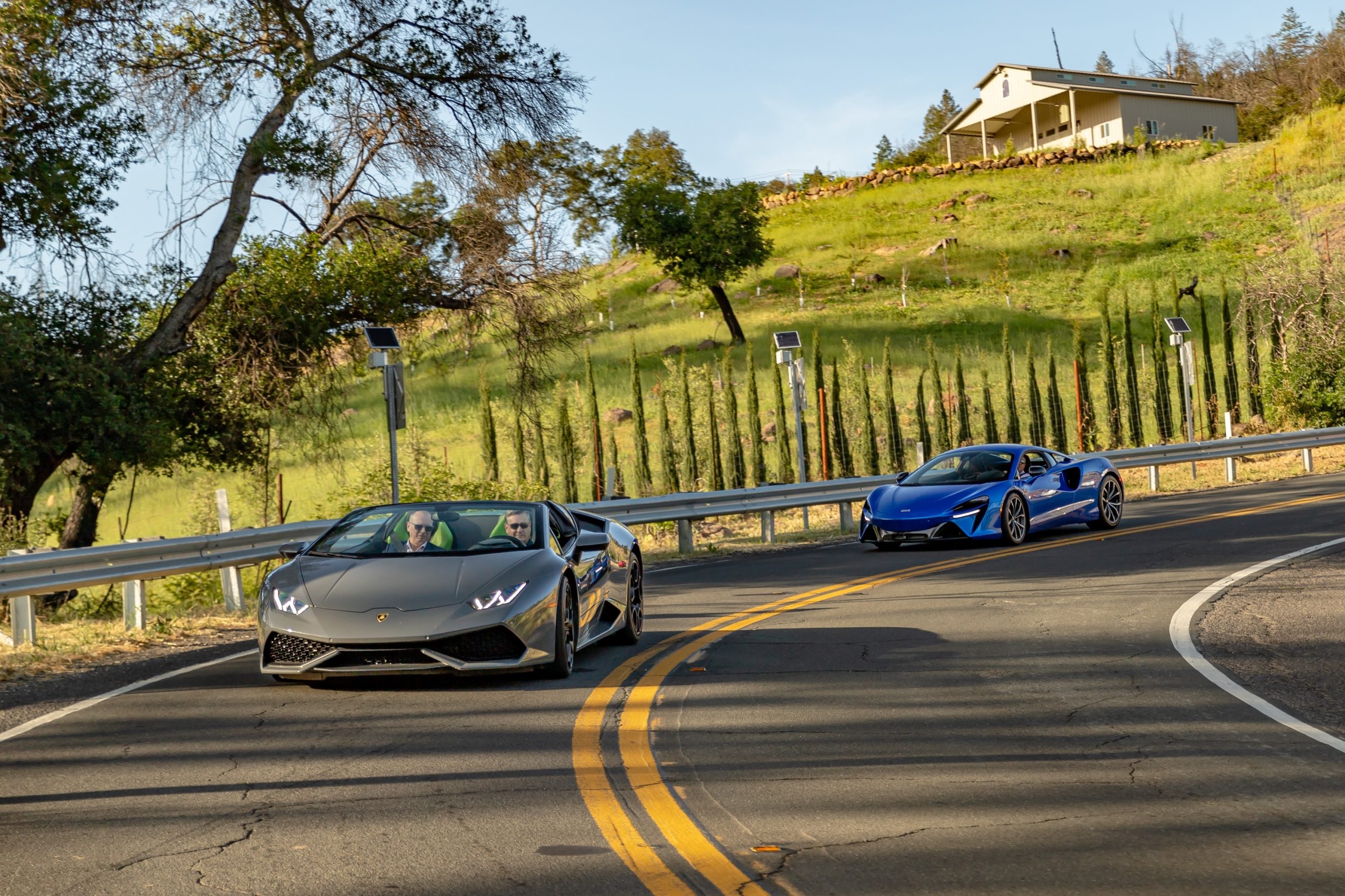 Luxury sports cars cruising on a winding road in Napa Valley, surrounded by lush greenery and scenic views.