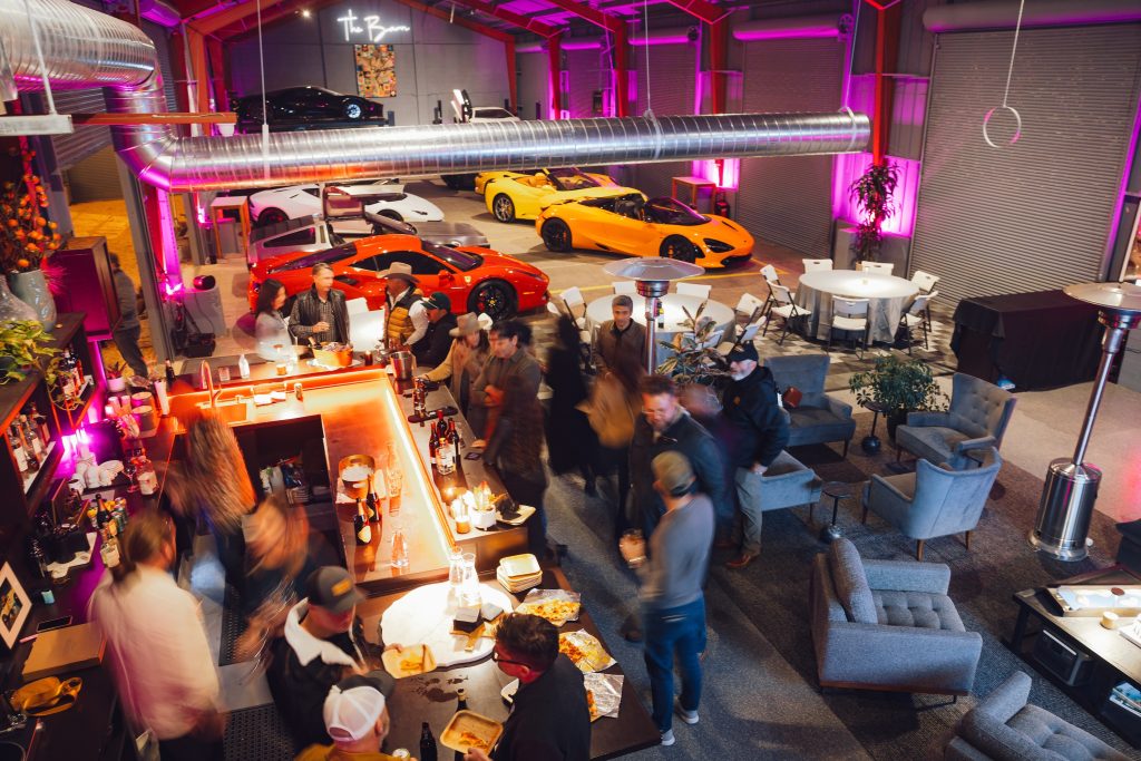 A lively gathering in a luxury venue featuring sports cars, a bar area, and guests enjoying food and drinks.