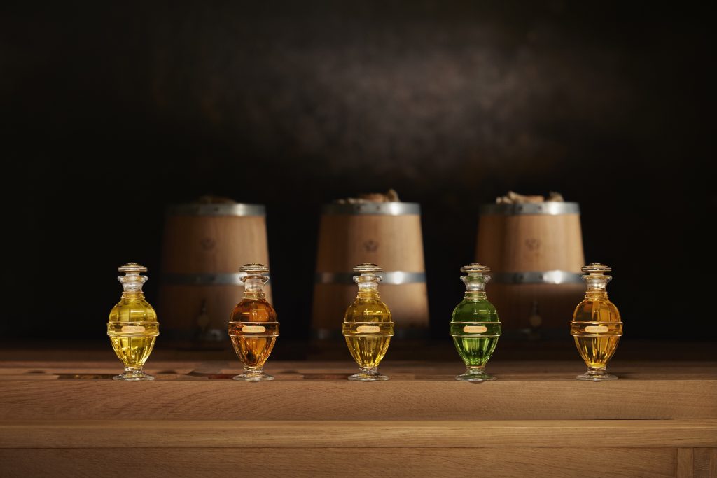 Five elegant glass perfume bottles in various colors are displayed against a dark background with wooden barrels.
