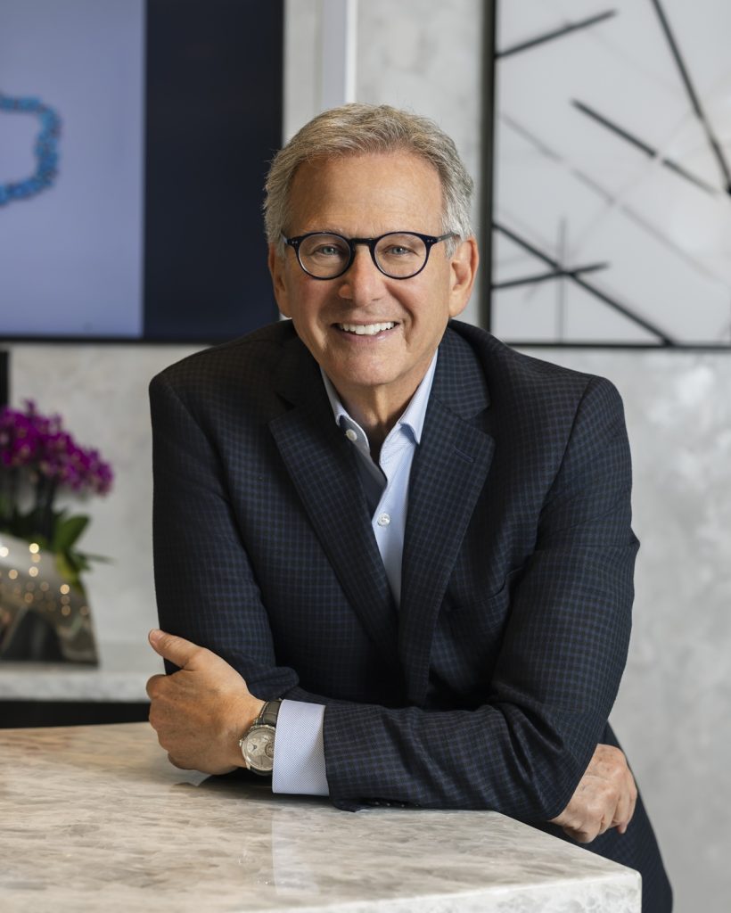 Martin Katz smiles while resting his arms on a marble countertop, dressed in a dark suit with a light blue shirt and glasses.