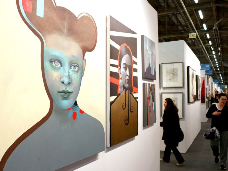 Artworks displayed on white walls, showcasing various styles and subjects, with visitors walking through the gallery.
