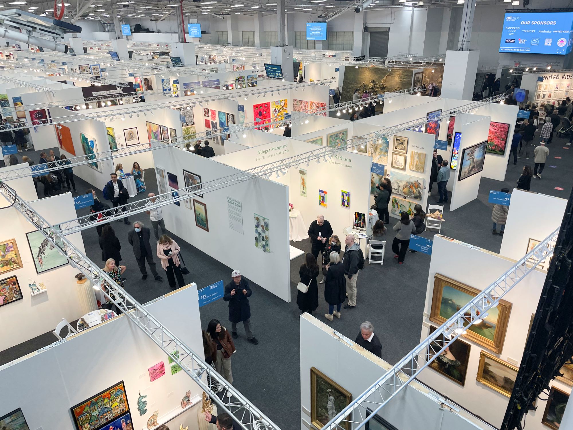 Crowd exploring various art booths filled with paintings at Artexpo New York, held at Pier 36 in Manhattan.