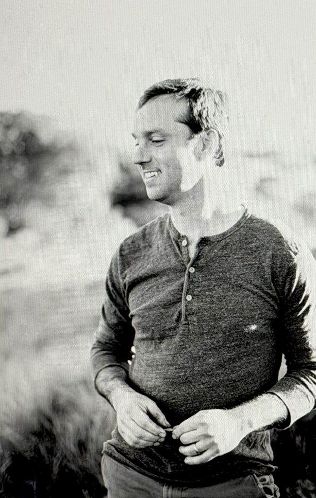 A man smiling and standing outdoors, wearing a gray henley shirt, surrounded by a natural landscape.