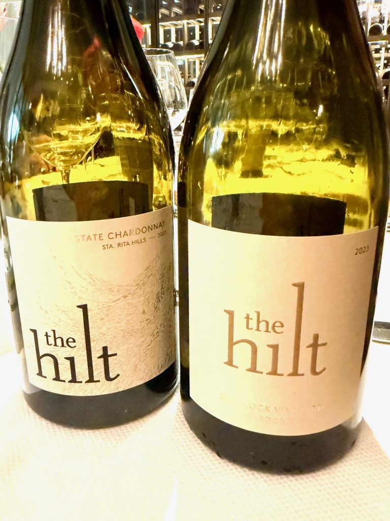 Two bottles of The Hilt wine, featuring Estate Chardonnay and Bentrock Vineyard labels, against a blurred background.