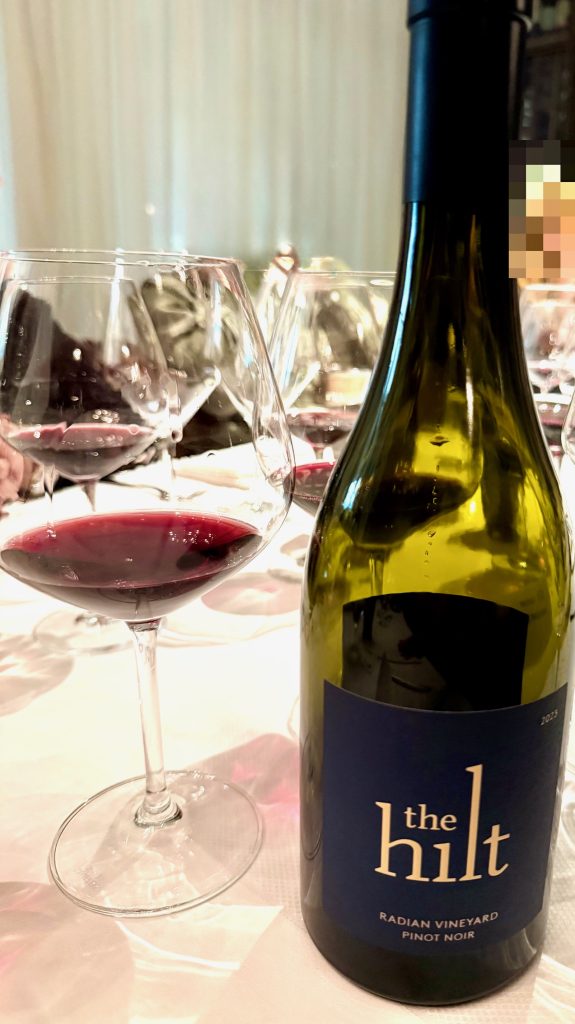 A bottle of The Hilt Radian Vineyard Pinot Noir beside a filled wine glass at a dining table setting.