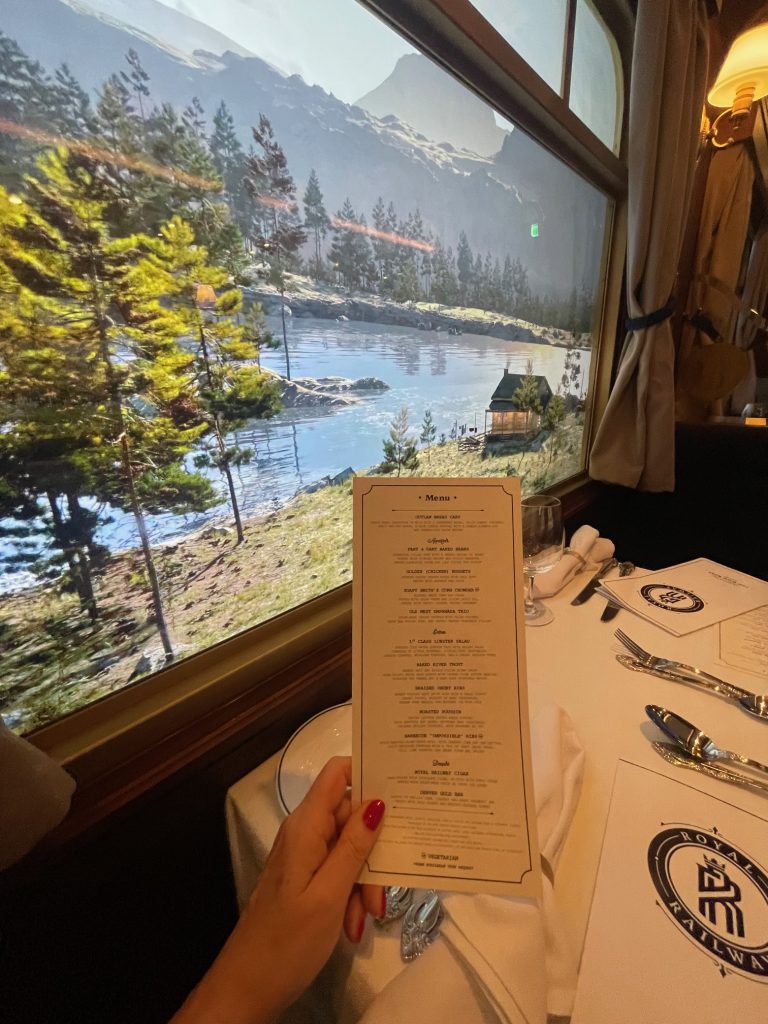 A hand holds a menu in front of a large window displaying a scenic view of mountains, forests, and a calm body of water.