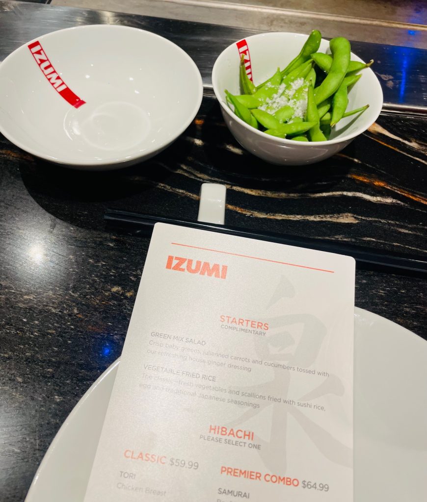White bowls with red "IZUMI" branding, one filled with edamame, and an open menu displaying Japanese starters and prices.