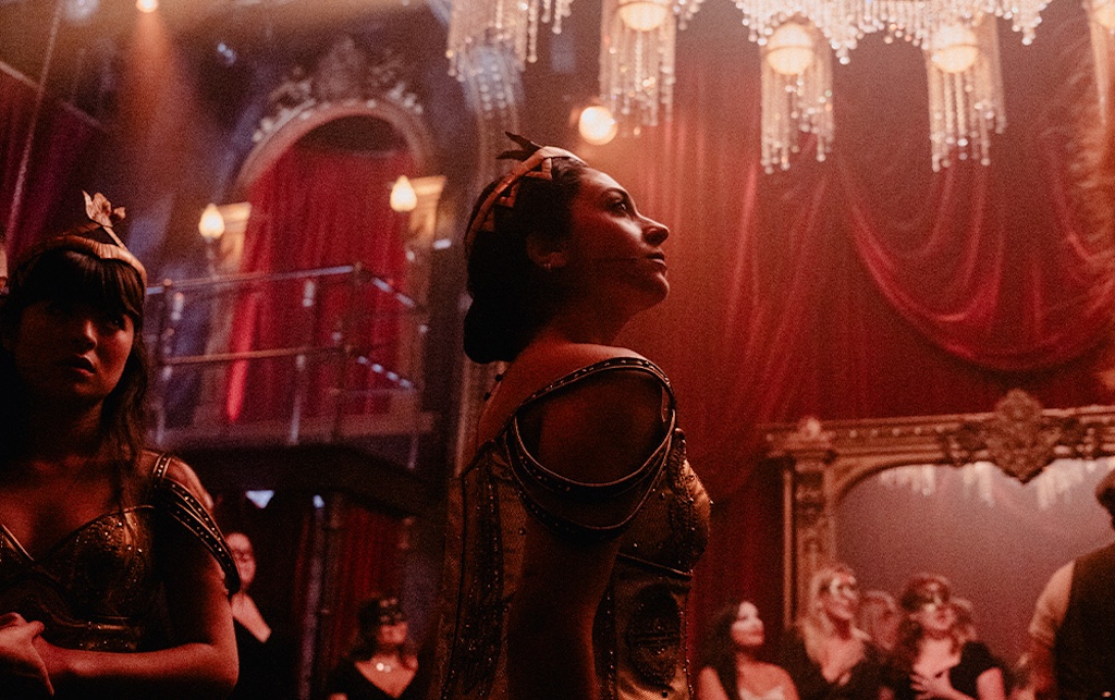 Two women in elaborate costumes stand in a dimly lit, ornate room adorned with red curtains and crystal chandeliers.