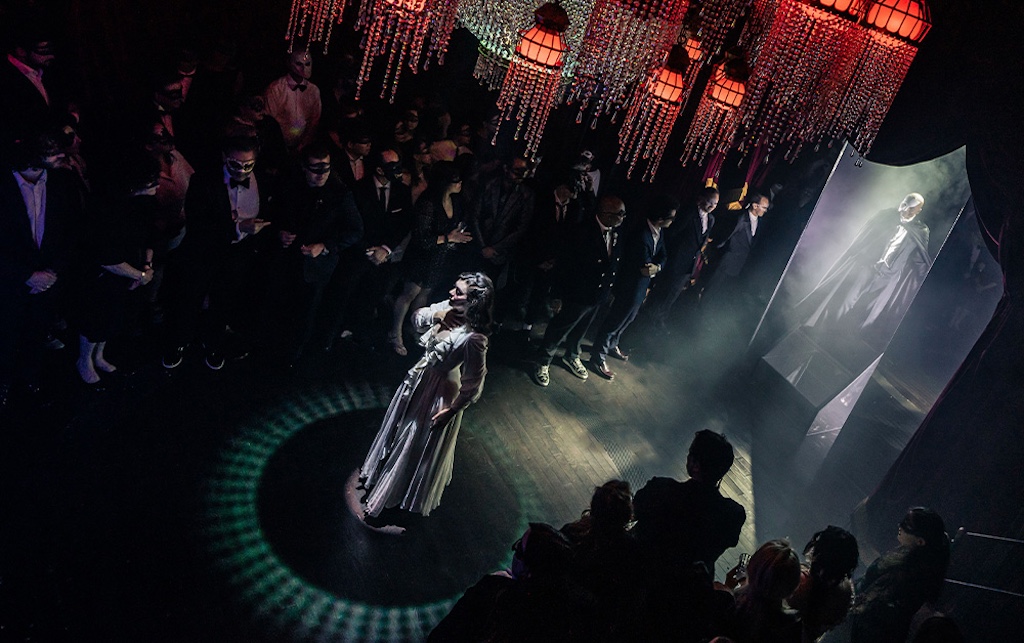 A performer in a white dress stands at the center of a dimly lit stage, surrounded by an attentive masked audience.