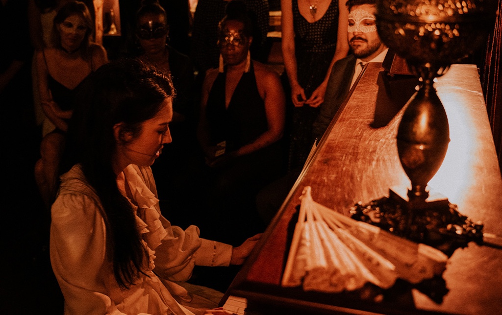 A woman plays the piano while an audience in masquerade masks watches intently in a dimly lit setting.