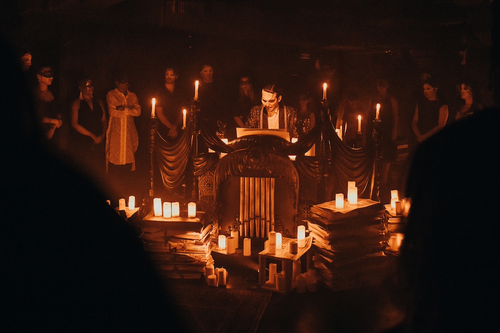 A performer reads in a dimly lit space surrounded by candles and an audience wearing masks, creating an atmospheric scene.