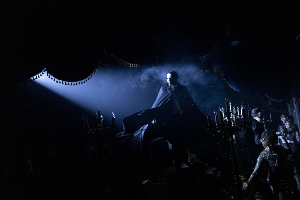 A figure in a dark cape stands under a spotlight, surrounded by a mysterious atmosphere and blurred silhouettes of other characters.