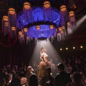 A performer in a gold dress stands under a chandelier, spotlighted by beams of light, while an audience in masks watches.