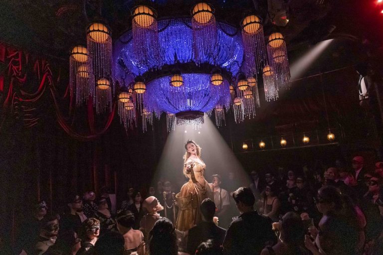 A performer in a gold dress stands under a chandelier, spotlighted by beams of light, while an audience in masks watches.