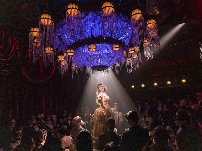 A performer in a gold dress stands under a chandelier, spotlighted by beams of light, while an audience in masks watches.