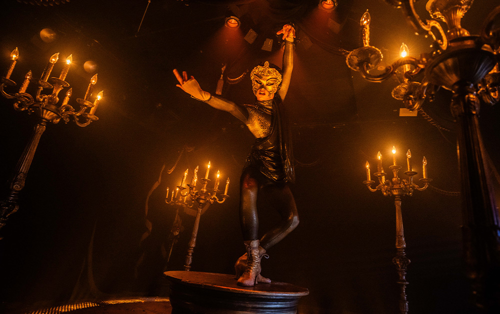 A performer in a mask and costume poses dramatically on a pedestal, surrounded by glowing chandeliers.